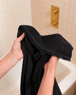 A close-up of a person holding a black bath towel with a fringed edge in front of a beige tiled bathroom wall and gold faucet.