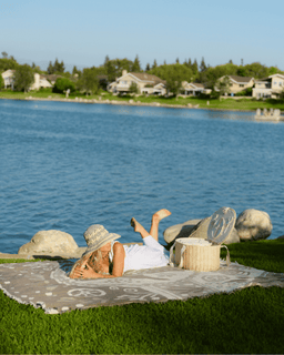 Woman relaxing on a blanket by a lake with a scenic view of houses and trees.
