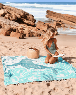 Woman sitting on a beach with a blue patterned towel, holding a book, and a woven bag nearby.