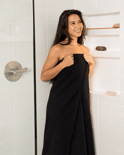 A woman wrapped in a black towel stands in a bright white tiled shower area, smiling and holding the towel around her body.