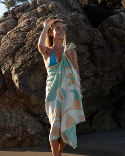 A person standing by a rock pool wearing a swimsuit and a towel with a tropical leaves design wrapped around the shoulders.
