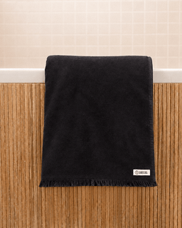 A black cotton bath towel with fringed edges hanging over a white bathtub edge, set against a wooden wainscoting and beige tiled wall background. A small white brand label reading 'SAND CLOUD' is visible in the bottom right corner of the towel.