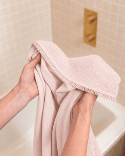 A person holding a soft blush pink bath towel with fringed edges in front of a tiled bathroom wall and gold wall-mounted fixture.