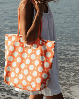 Person holding an orange tote bag with white polka dots on a beach.