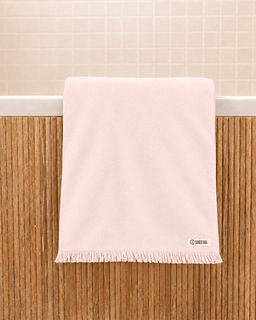 A pale pink bath towel with fringe trim hanging over the edge of a bathtub against a bamboo screen and tiled wall.