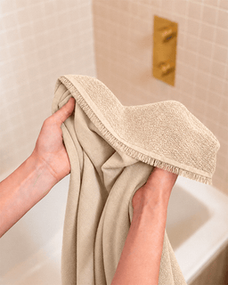 A person holding a beige textured bath towel with fringed edges in front of a tiled bathroom wall and white bathtub.