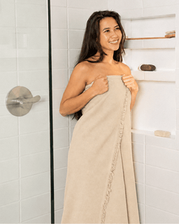 A woman wrapped in a beige towel stands in a bright white tiled shower area with built-in shelves.