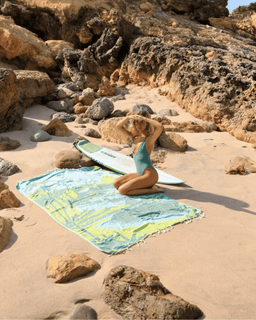 Person in a blue swimsuit sitting on a colorful towel with a surfboard next to them on a rocky beach.