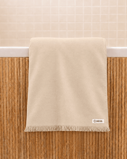 A beige cotton bath towel with fringe trim hangs over the edge of a bathtub against a wooden wainscoting and tiled wall background. A small white brand label reading 'SAND CLOUD' is visible near the bottom right corner of the towel.