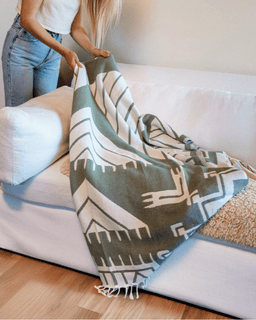 Person holding a patterned green and beige blanket over a white couch.
