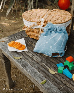 Wicker basket, blue drawstring bag, orange slices, and colorful blocks on a wooden surface outdoors.