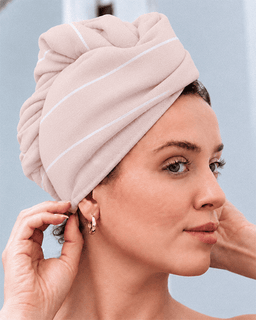 A woman wearing a soft, twisted bath towel turban hat in a dusty pink color with thin white stripes, adjusting it on her head.