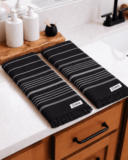Two black towels with white stripes and fringe, laid out on a white bathroom countertop next to a sink.