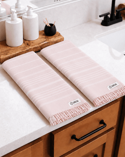 Two pale blush pink kitchen towels with thin white stripes and fringed edges, laid on a white countertop in a bathroom setting.
