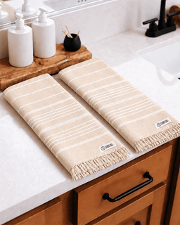 Two beige and white striped towels with fringe edges laid out on a white bathroom countertop next to a sink.
