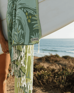 Person holding a surfboard with a green towel featuring a palm tree design, beach in the background