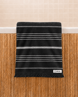 A black cotton bath towel with horizontal white stripes and fringed edges, hanging on a bamboo wall rack in front of a beige tiled wall.
