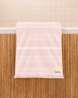 A light pink cotton bath towel with thin white horizontal stripes and a fringed edge, draped over the side of a bathtub with a beige tiled wall in the background.