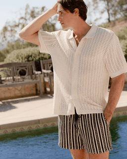 Man wearing a light-colored knit shirt and striped shorts by a poolside.