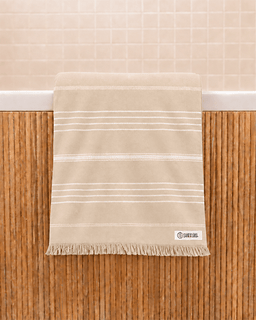 A beige cotton bath towel with thin white horizontal stripes and a fringed hem, hanging over a white tub edge against a bamboo screen and tiled wall.