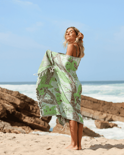 Woman wrapped in a green patterned towel on a beach with ocean and rocks in the background