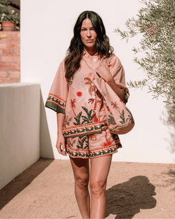 Woman wearing a pink embroidered dress standing outdoors with a white wall and greenery in the background.