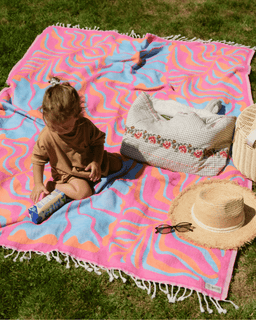 Child sitting on a colorful blanket with a bag and hat on a grassy background