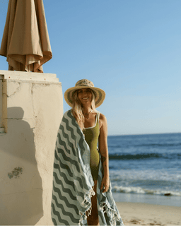 Woman on a beach with a towel draped over her, standing near an umbrella.