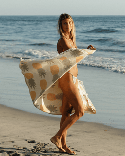 Woman on a beach holding a pineapple-patterned towel with ocean waves in the background