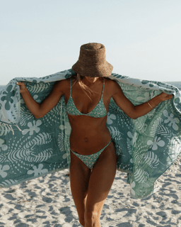 Woman in a green bikini with a patterned towel and straw hat on a beach.