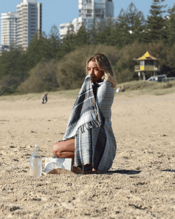 Woman sitting on a sandy beach with a plaid blanket, surrounded by trees and buildings.