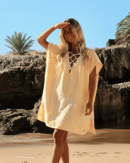 Woman in a beige cover-up standing on a beach with rocky cliffs and palm trees in the background.