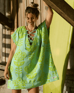 Woman wearing a green and blue floral dress standing next to a yellow surfboard.
