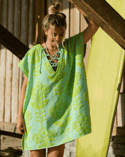 Woman wearing a green and yellow patterned dress standing next to a yellow surfboard.