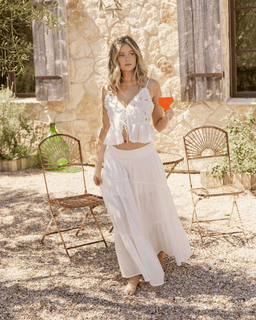 Woman in a white dress holding a drink outdoors near stone walls and metal chairs.