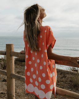 Woman in an orange dress with white polka dots standing by a wooden railing overlooking the ocean.