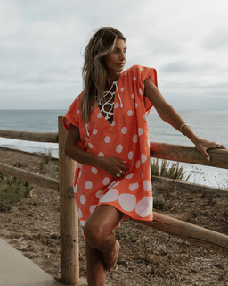 Woman in an orange polka dot dress standing by a wooden railing with ocean in the background