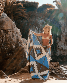 Woman holding a patterned blanket on a rocky beach with palm trees in the background