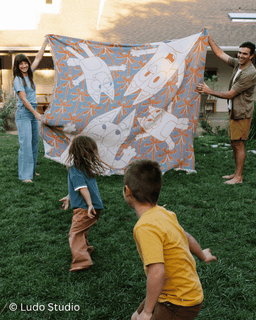 Family playing with a colorful blanket featuring cartoon characters on a grassy lawn.