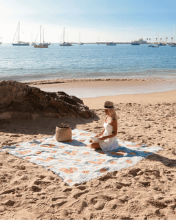 Woman sitting on a floral beach blanket with a hat, surrounded by boats in the water.