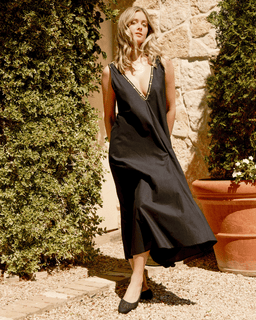 Woman in a black dress standing outdoors with greenery and stone wall in the background