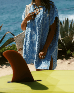 Person wrapped in a blue patterned towel by the ocean with a surfboard in the foreground.