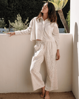 Woman in a white outfit standing against a white wall with greenery in the background