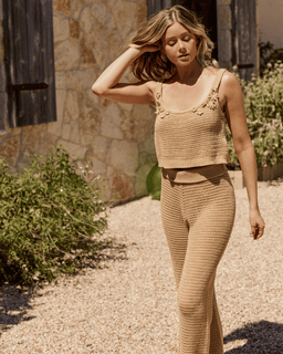 Woman wearing a beige crochet outfit standing outdoors near a stone wall.