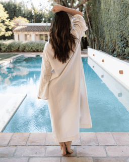 Woman in a long, light-colored robe standing by a pool with a house and trees in the background.
