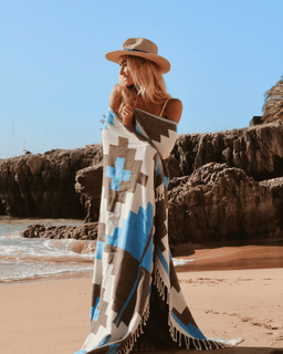 Woman on a beach holding a patterned blanket with rocky cliffs and ocean in the background