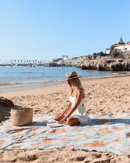 Woman sitting on a floral blanket on a beach with ocean and buildings in the background