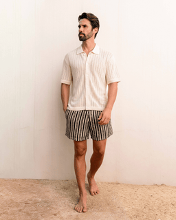 Man wearing a white textured shirt and patterned shorts standing against a plain background