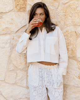 Woman in a white outfit drinking from a glass against a stone wall