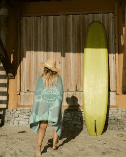 Woman in a blue towel and straw hat standing next to a green surfboard against a wooden wall.